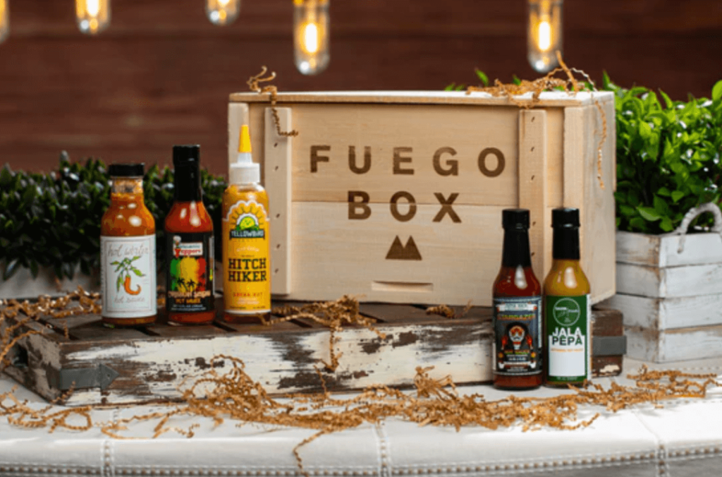 A wooden box that says "Fuego Box" surrounded by 5 different types of hot sauces.